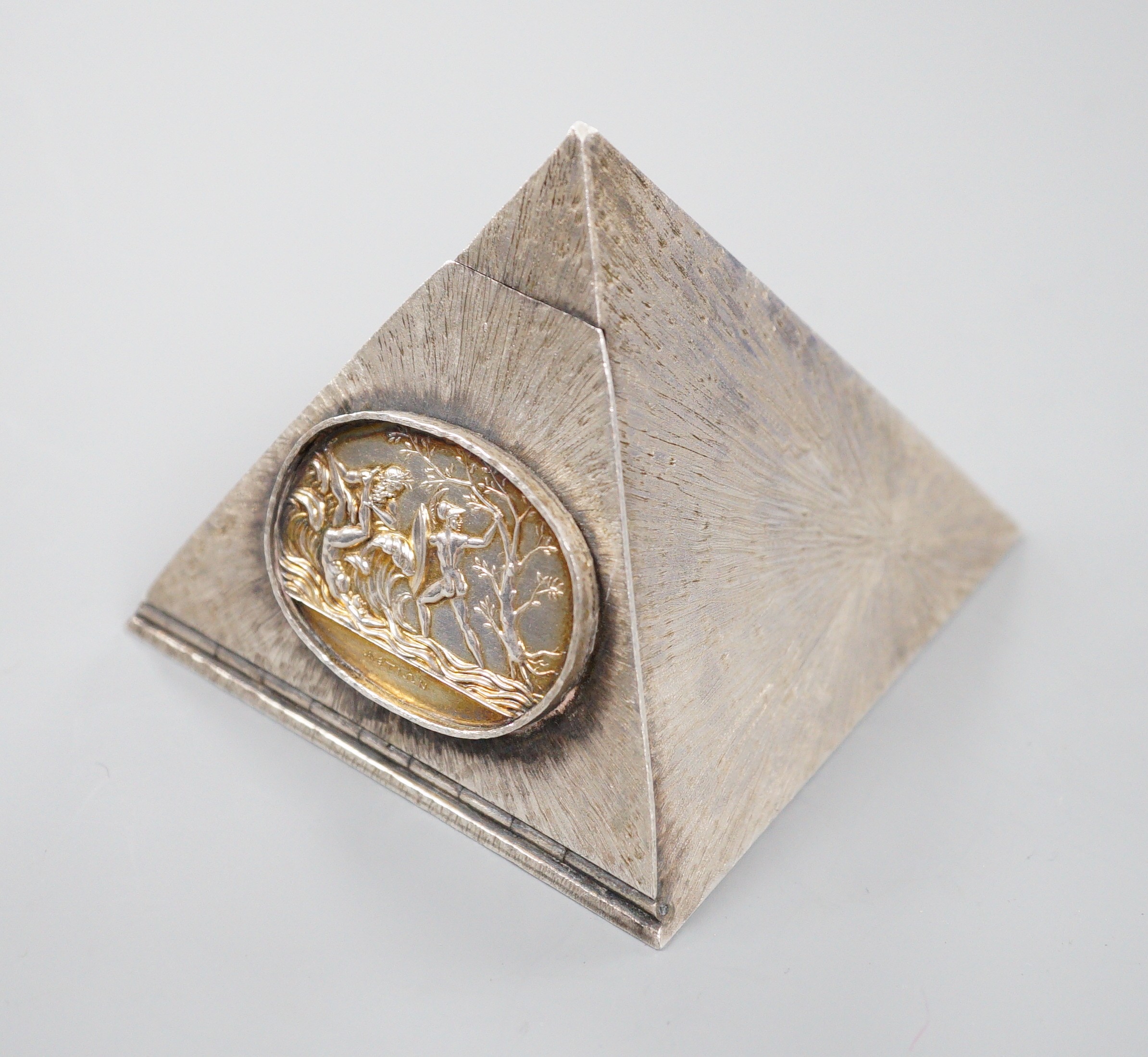 A textured white metal pyramid shaped box, the hinged panel decorated with Greek scene and containing five dice, height 62mm.